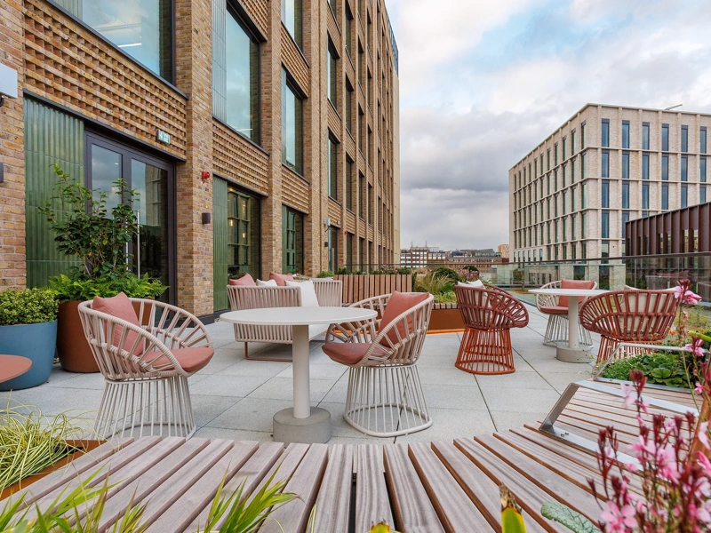 A spacious balcony at One Blossom Yard, UK, features chairs and a small table with potted plants. The skyline and distant mountains create a scenic backdrop against a cloudy afternoon sky.