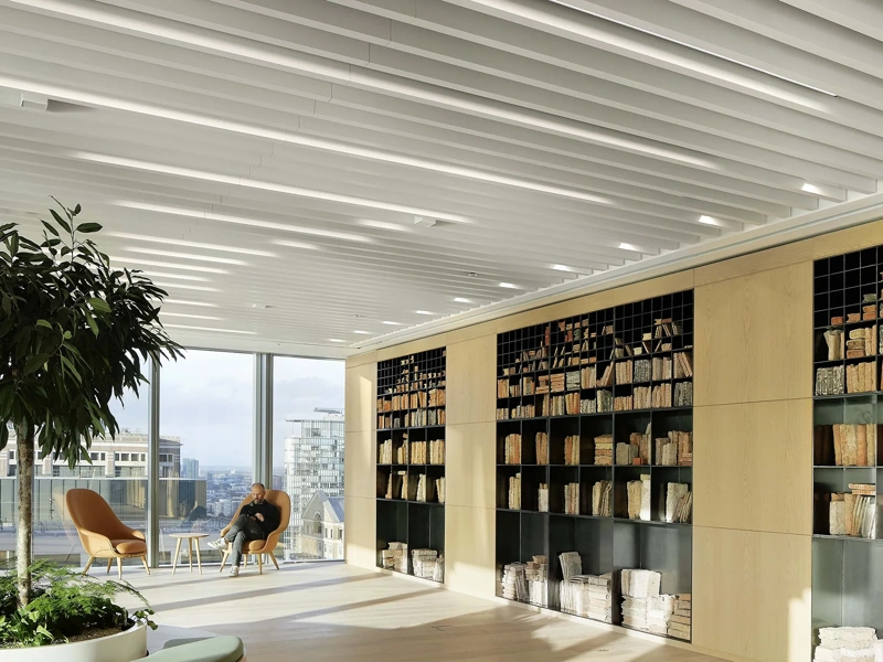 Bright plant filled office space with glass windows and shelf full of books. There's a man in the corner on a orange chair.