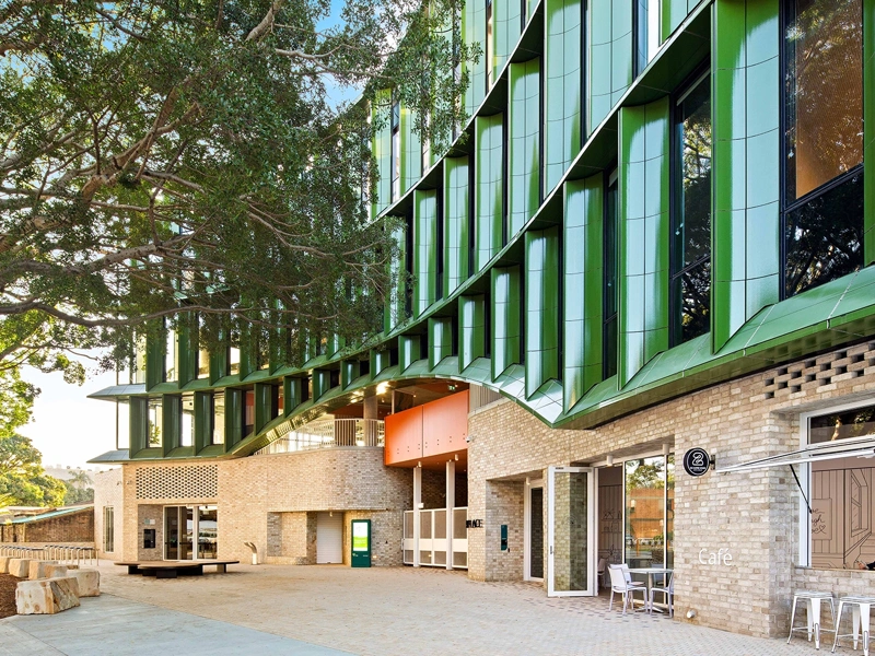 The exterior of a modern, green building with foliage and seating next to it.