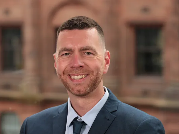 Portrait of Mark Ferguson, Associate, UK, standing against brown building in blue suit.