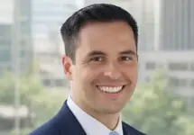 Portrait of Andrew Pollard, Director, standing against office window with tall city views behind and green trees in background.