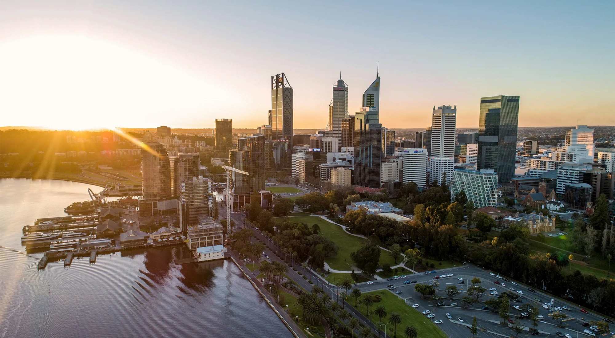 Aerial view of modern city skyline at sunset, with high rise buildings, a riverfront with docks and green parkland in the foreground.