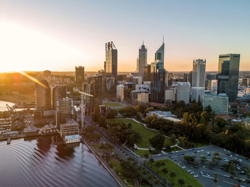 Aerial view of modern city skyline at sunset, with high rise buildings, a riverfront with docks and green parkland in the foreground.