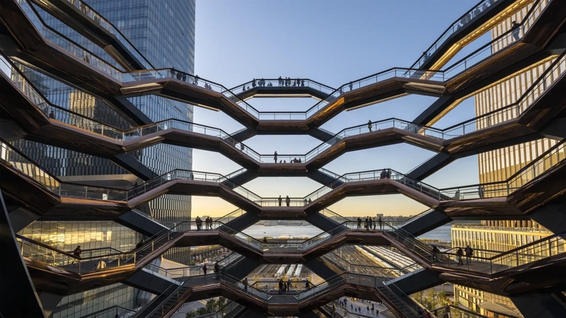 Architectural view of the Vessel at Hudson Yards in New York City, showing multiple levels of interconnected staircases with silhouetted people walking against a backdrop of modern skyscrapers and a sunset sky