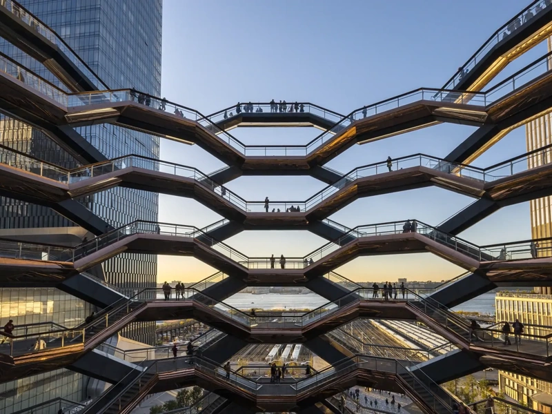 Architectural view of the Vessel at Hudson Yards in New York City, showing multiple levels of interconnected staircases with silhouetted people walking against a backdrop of modern skyscrapers and a sunset sky