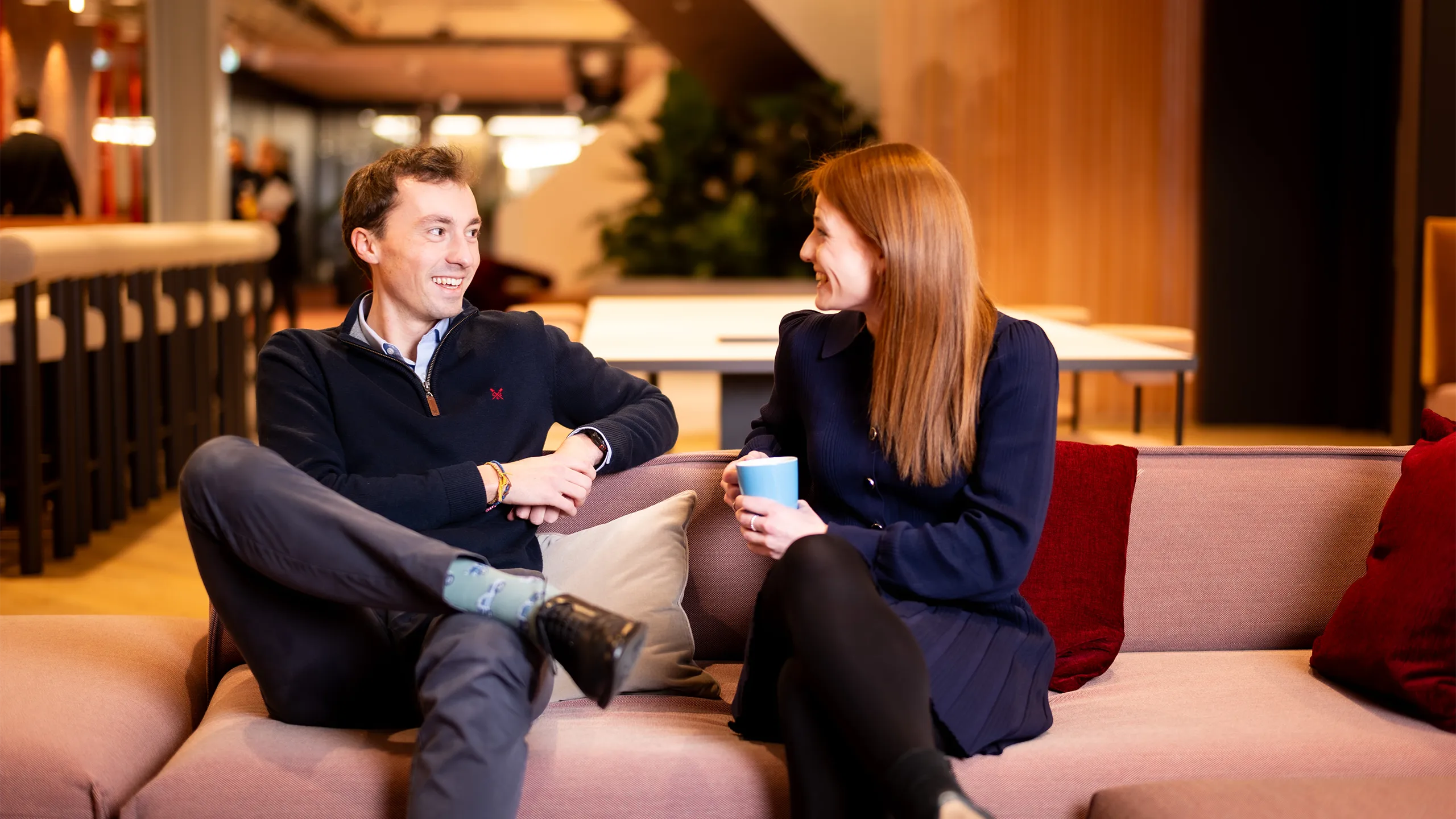 Male and female colleagues sitting together on light pink sofas in break out room having a casual chat.