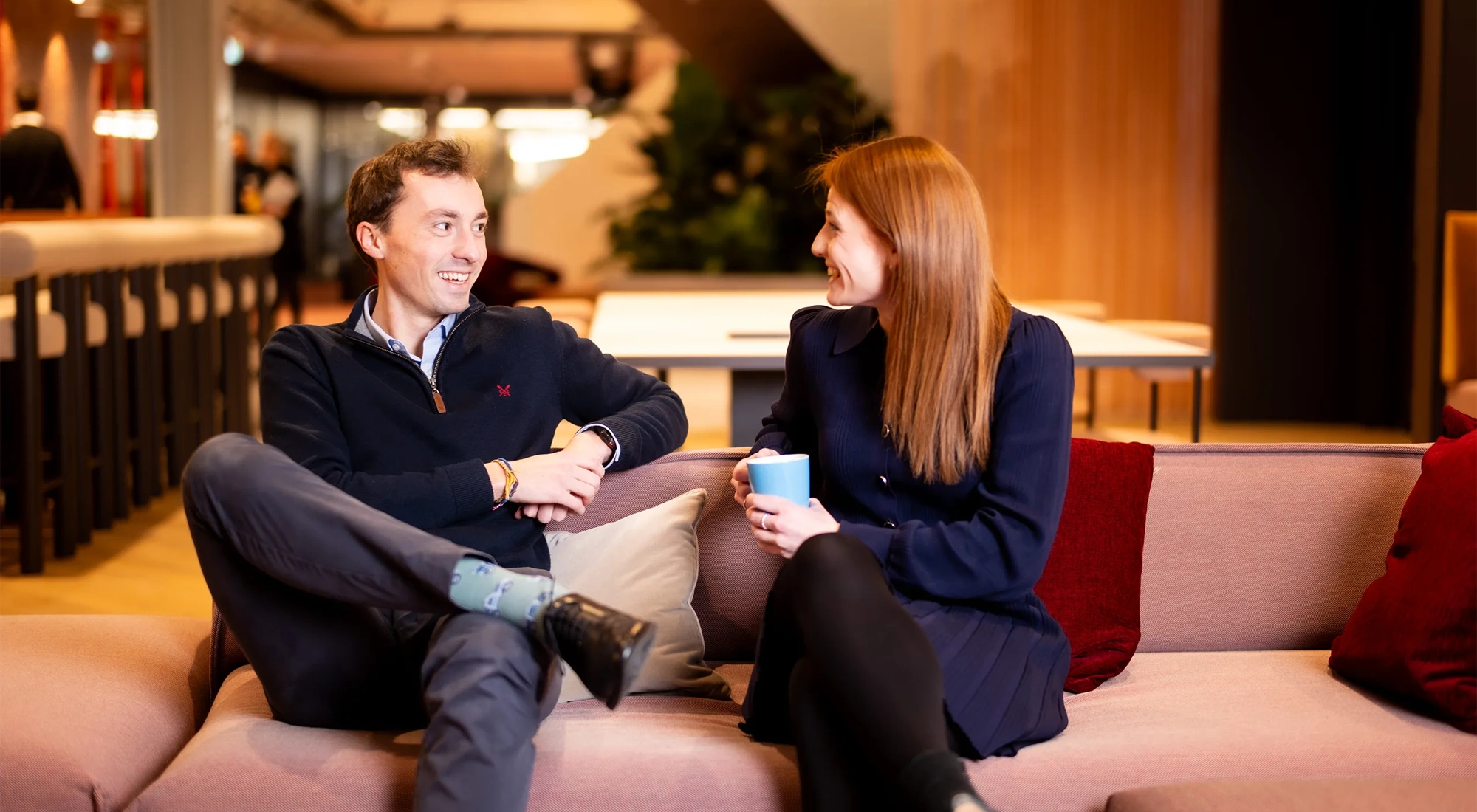 Male and female colleagues sitting together on light pink sofas in break out room having a casual chat.