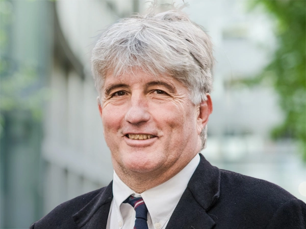 Mark Wainwright, Managing Director, Mining, standing outside in front of an outdoor urban setting, featuring modern architectural elements with clean lines. There are hints of greenery, suggesting some trees or plants.
