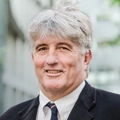 Mark Wainwright, Managing Director, Mining, standing outside in front of an outdoor urban setting, featuring modern architectural elements with clean lines. There are hints of greenery, suggesting some trees or plants.