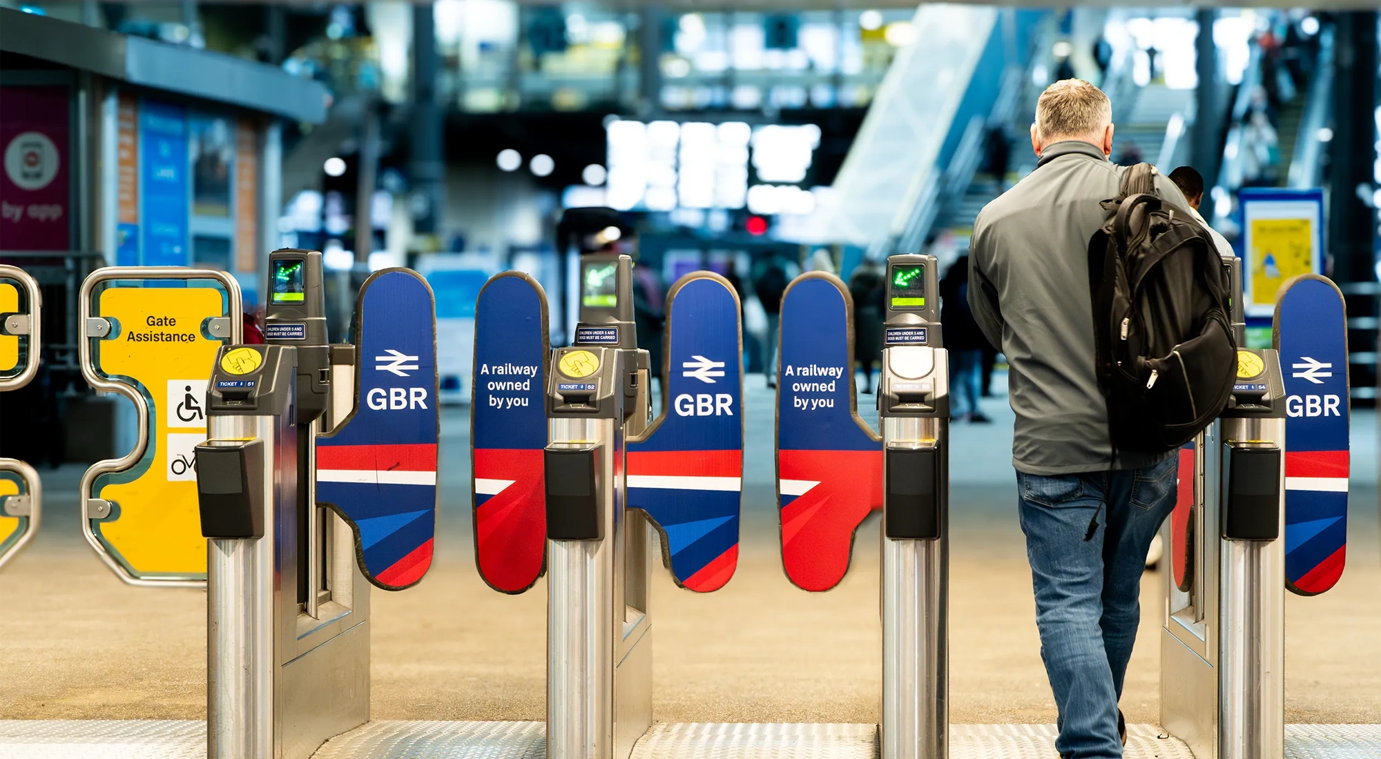 A rail passenger going through the ticket barrier at Leeds Train Station with the UK Government's re-nationalised rail company GBR signs and symbols displayed
