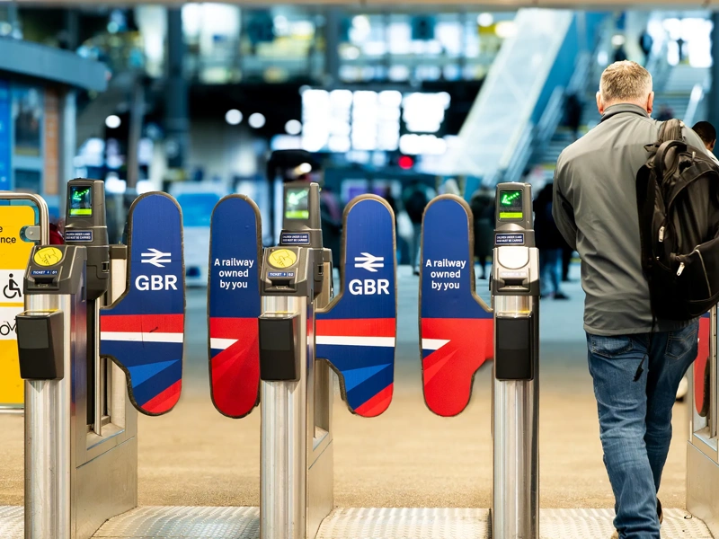 A rail passenger going through the ticket barrier at Leeds Train Station with the UK Government's re-nationalised rail company GBR signs and symbols displayed