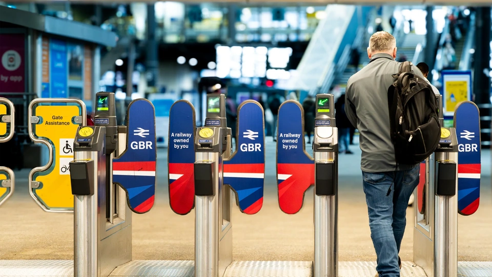 A rail passenger going through the ticket barrier at Leeds Train Station with the UK Government's re-nationalised rail company GBR signs and symbols displayed