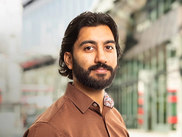 Muntasir Hussein standing outside in front of buildings featuring an urban cityscape with a mix of architectural styles. The scene includes some windows and red features, adding a touch of color.