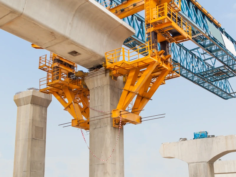 A large yellow and blue bridge construction crane is positioned above concrete support pillars, actively building a segmental bridge. The structure shows a partially completed bridge span with support equipment and materials on the site