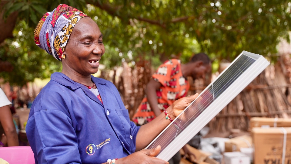 Woman wearing a purple Barefoot College International shirt holding a solar panel outside, in front of trees