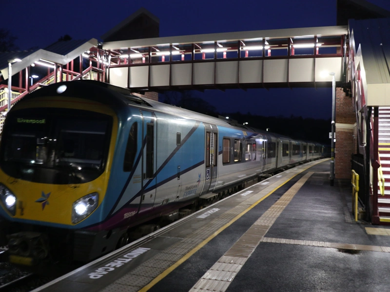 Photo of a passenger train passing under a pedestrian bridge that spans across platforms at a train station