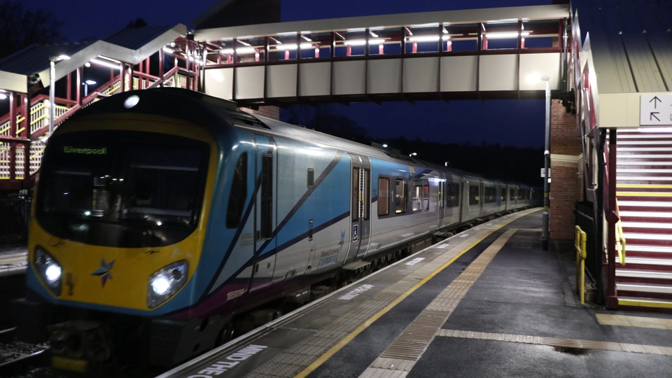 Photo of a passenger train passing under a pedestrian bridge that spans across platforms at a train station