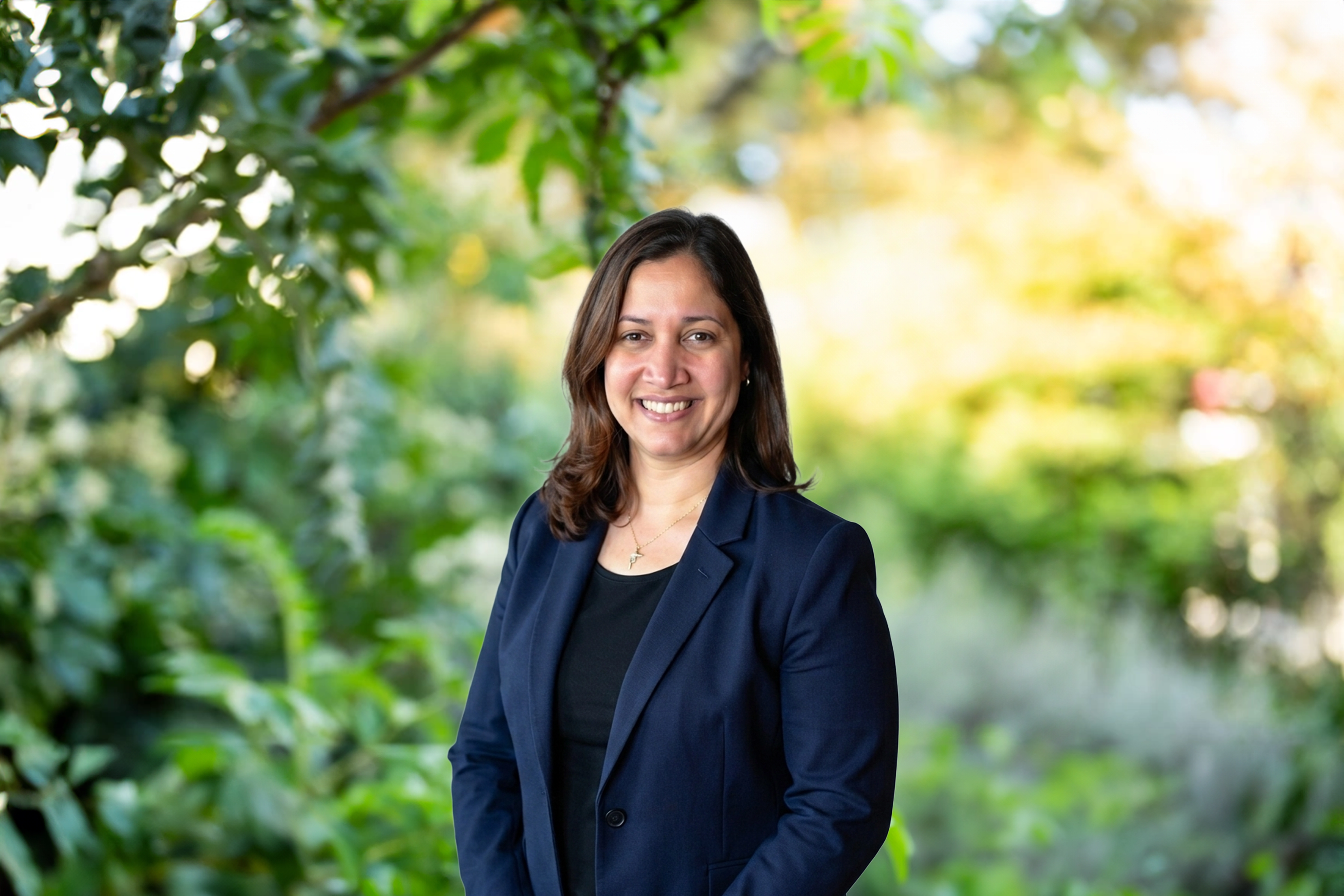 Indhira Figuereo standing outdoors in front of dense green foliage and landscaped bushes, with background softly blurred.