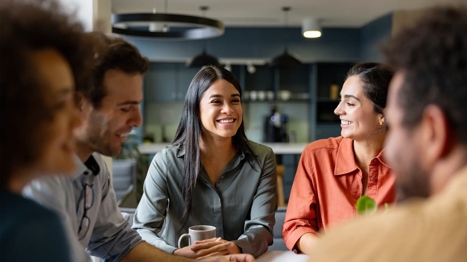 Group of colleagues sitting together having a conversation, gathered around a table with coffee mugs.