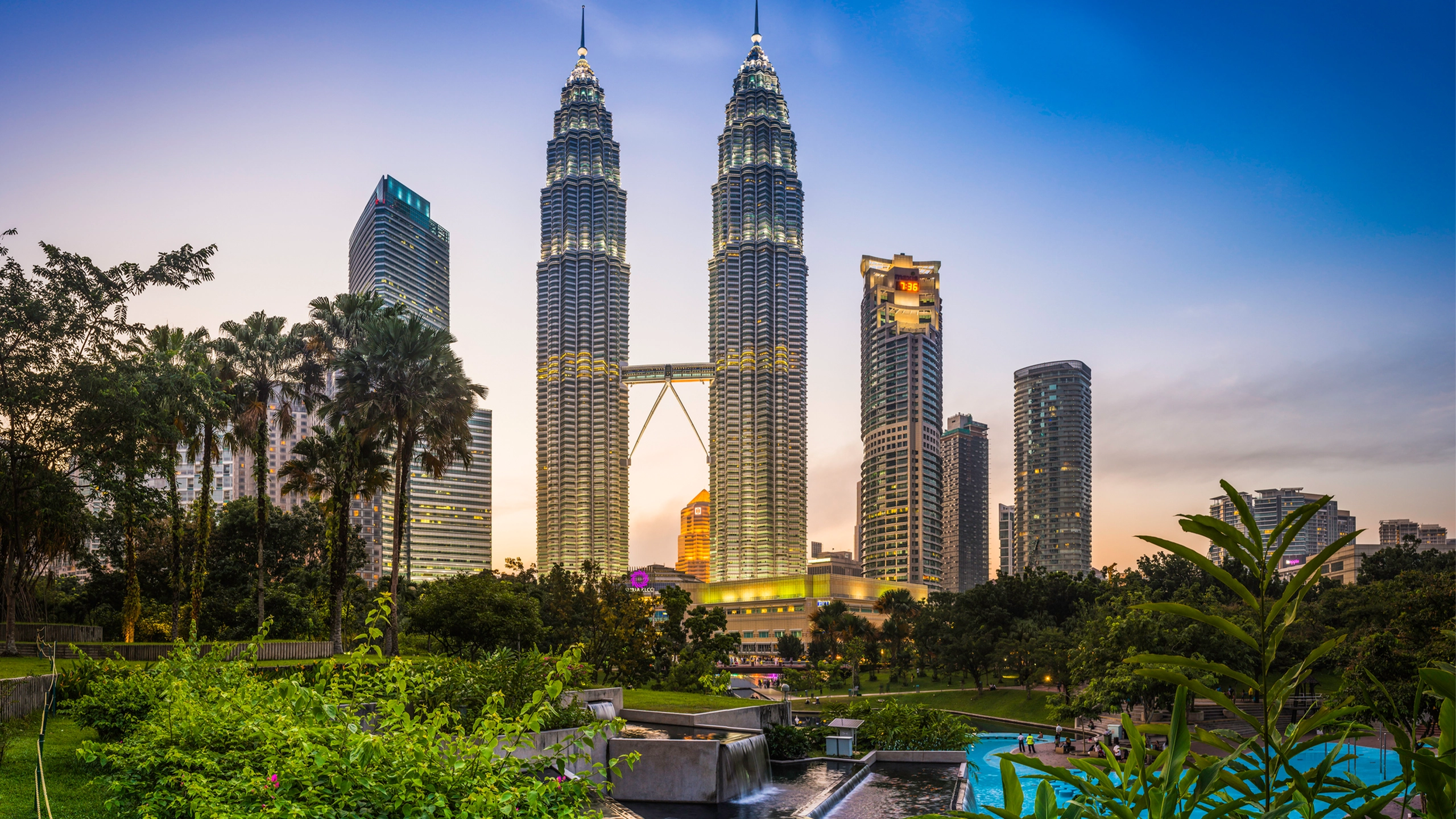 The iconic twin spires of the Petronas Towers glittering against the blue dusk sky above the ponds of KLCC Park and Suria KLCC in the heart of Kuala Lumpur, Malaysia's vibrant capital city