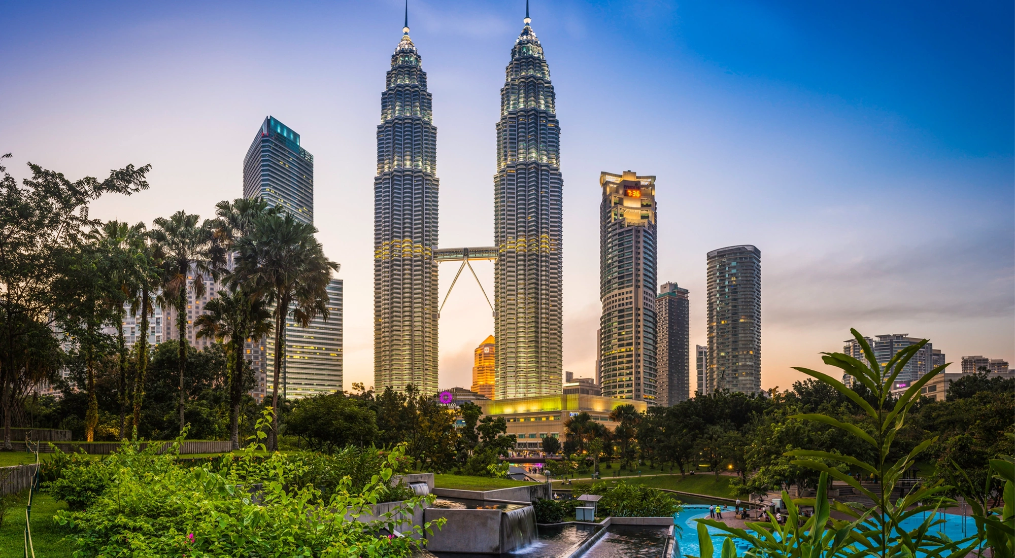 The iconic twin spires of the Petronas Towers glittering against the blue dusk sky above the ponds of KLCC Park and Suria KLCC in the heart of Kuala Lumpur, Malaysia's vibrant capital city