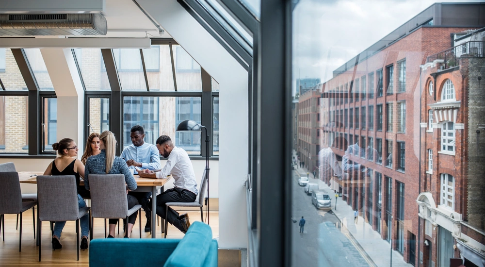 A group of male and female work colleagues sitting at a boardroom desk with exterior street view to the left
