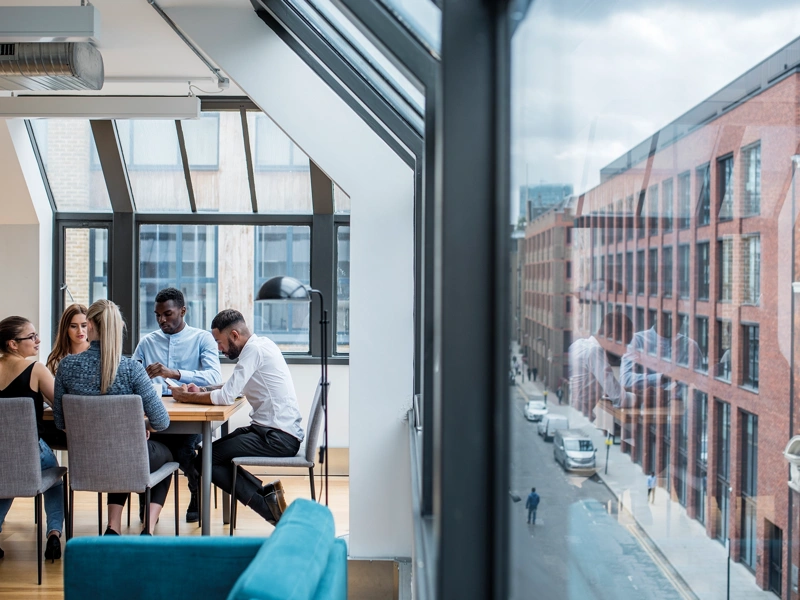 A group of male and female work colleagues sitting at a boardroom desk with exterior street view to the left