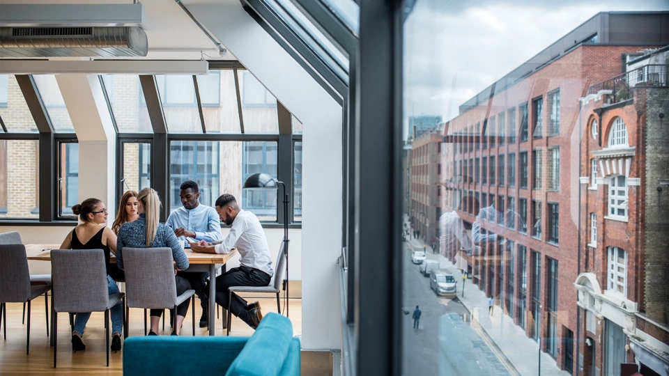 A group of male and female work colleagues sitting at a boardroom desk with exterior street view to the left