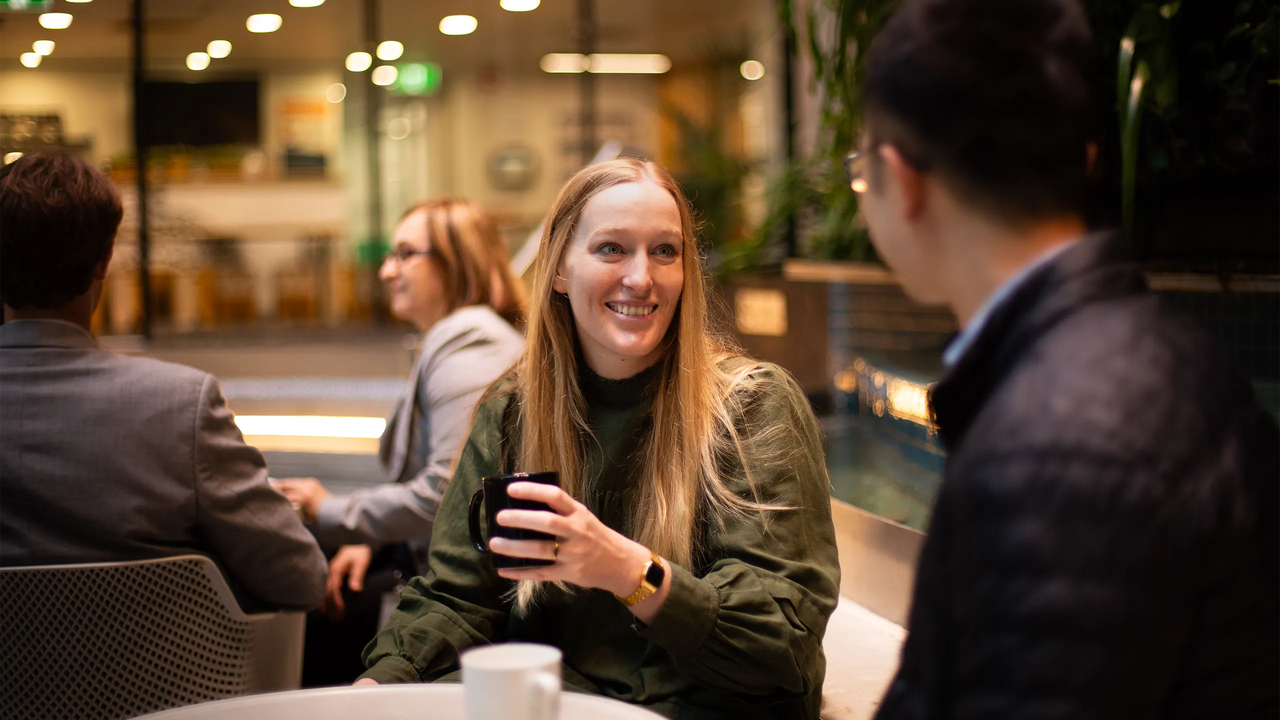 Two colleagues together shown from the males perspective, having a conversation in a plant-filled office, with blonde female colleague featured and smiling.