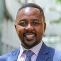 Robert Gichohi, Country Director, standing outside in front of an outdoor urban setting, featuring modern architectural elements with clean lines. There are hints of greenery, suggesting some trees or plants.