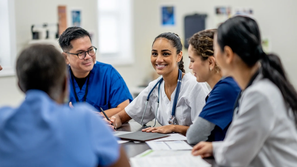 A small medical staff team sit around a boardroom table as they meet to discuss and collaborate on patient cases. They are each dressed professionally and are focused on the meeting.