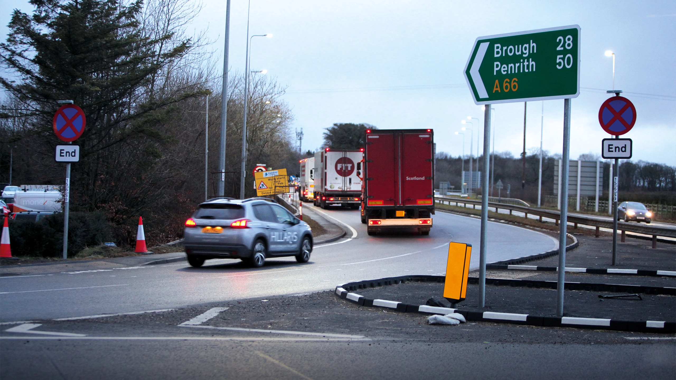 Photo of a car and two trucks joining an offramp on a road with sign on the right of the image indicating A66 route