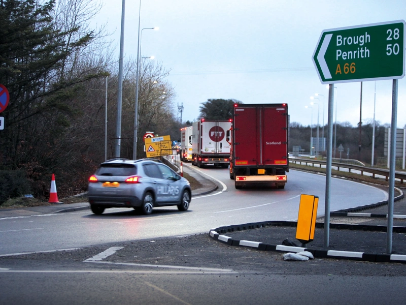 Photo of a car and two trucks joining an offramp on a road with sign on the right of the image indicating A66 route