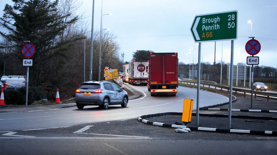 Photo of a car and two trucks joining an offramp on a road with sign on the right of the image indicating A66 route