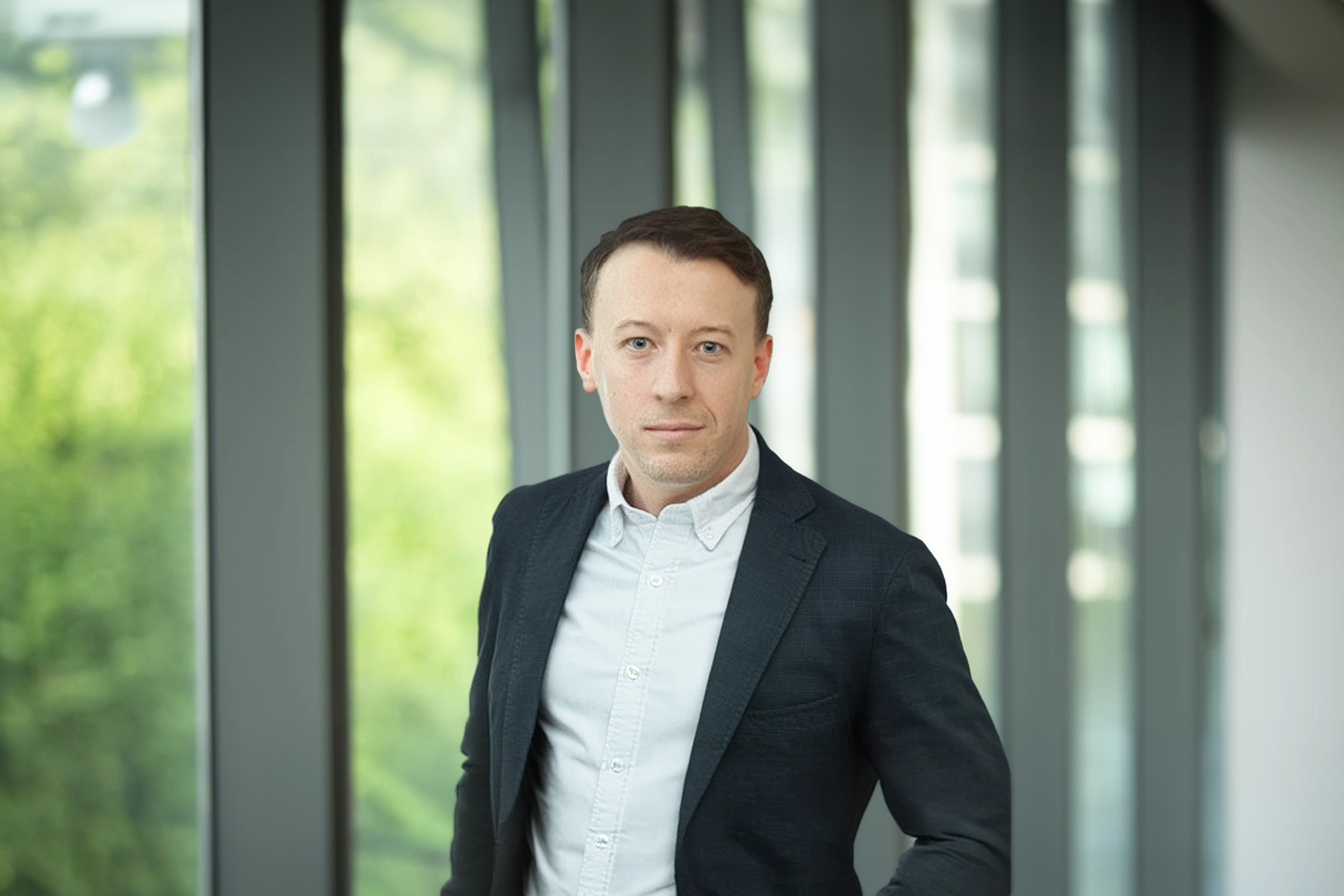 Oliver Molloy, standing in office in front of window showcasing an outdoor urban setting, featuring modern architectural elements with clean lines. There are hints of greenery, suggesting some trees or plants.