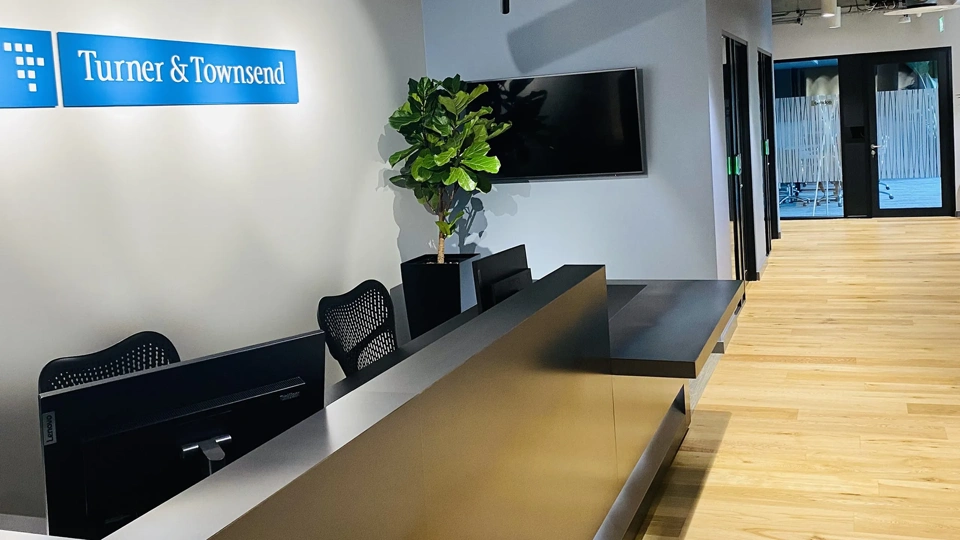 Picture of office reception and hallway with the sign 'Turner & Townsend' above reception chairs. There is a plant in the corner in front of a tv.