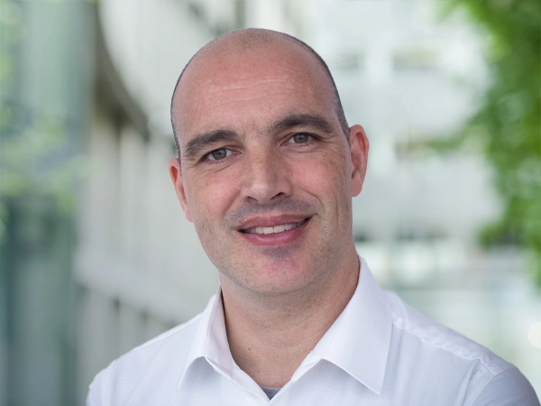 Hans Weemaes, Director, Sustainability, Asia, standing outside in front of an outdoor urban setting, featuring modern architectural elements with clean lines. There are hints of greenery, suggesting some trees or plants.