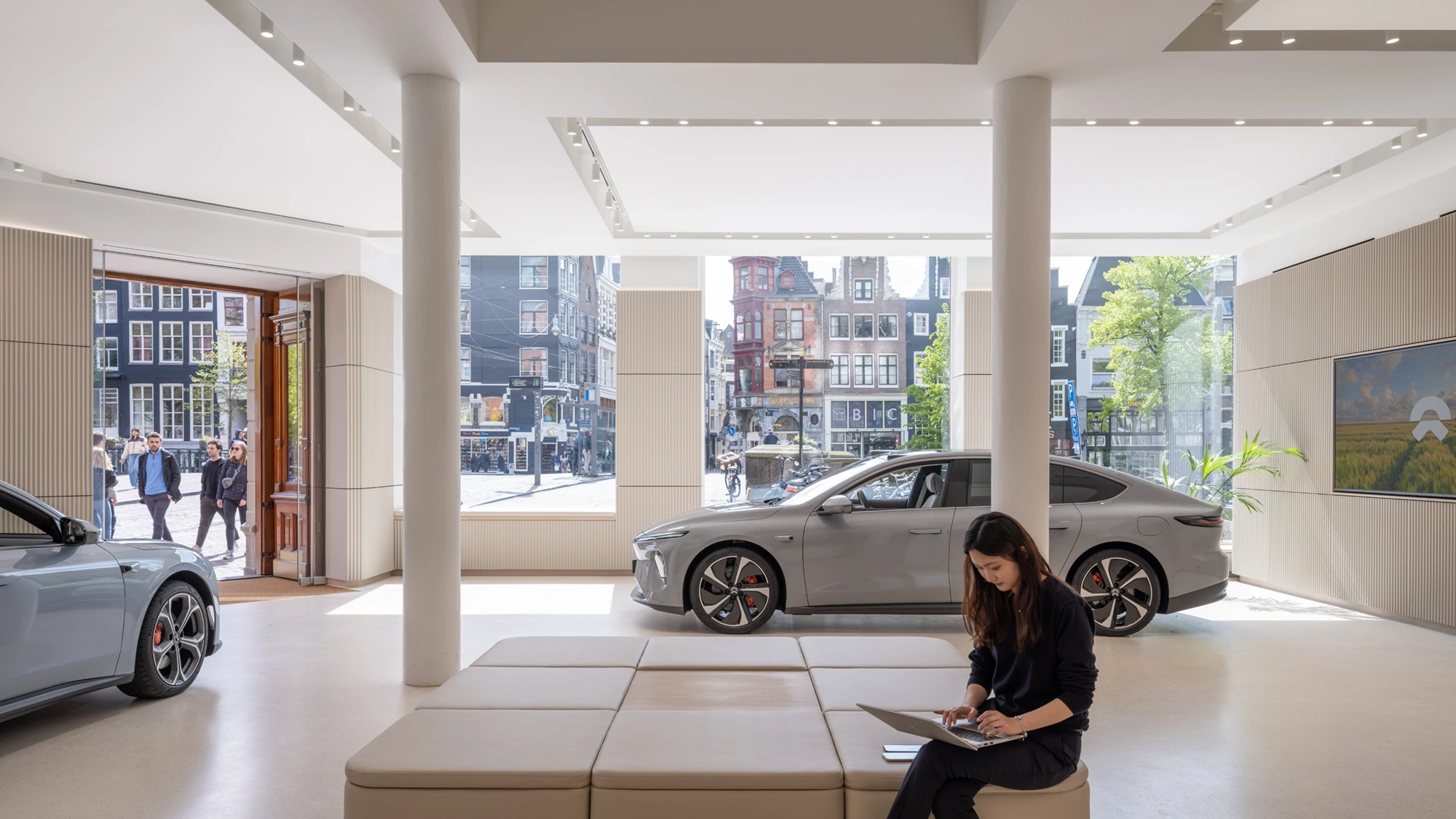 Interior of NIO gallery ground floor showroom. Two cars and a woman at a laptop feature in front of a glass backdrop of Amsterdam