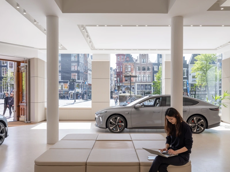 Interior of NIO gallery ground floor showroom. Two cars and a woman at a laptop feature in front of a glass backdrop of Amsterdam