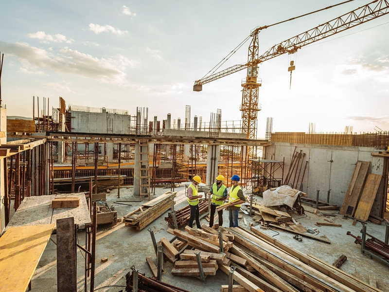 Three male construction workers discussing plans on the floor of a building under construction with a crane in background skyline.