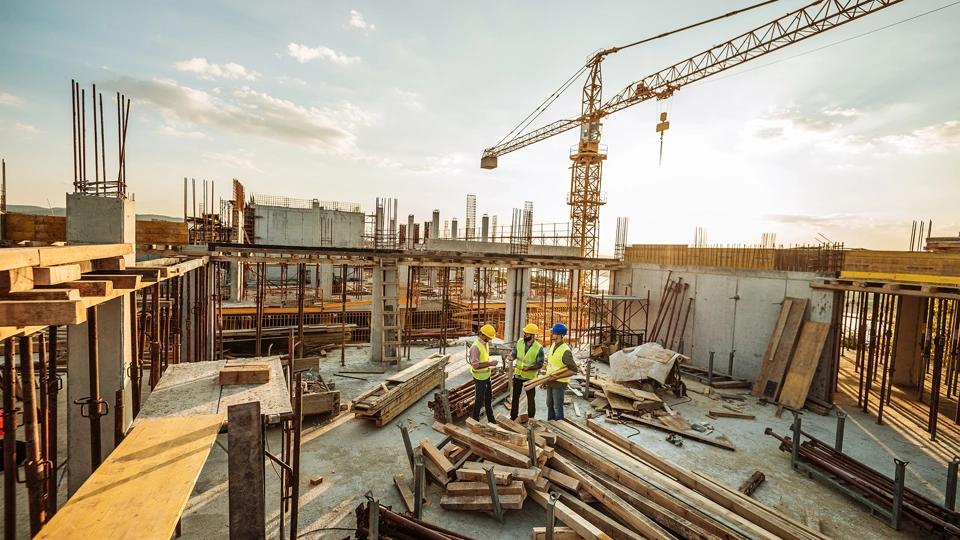 Three male construction workers discussing plans on the floor of a building under construction with a crane in background skyline.