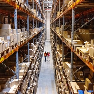 Two people, on in a safety vest, walking through a warehouse lined with cardboard boxes