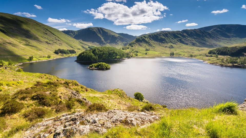Haweswater Reservoir in Mardale Valley taken from Whiteacre Crag. A green hill that leads down to the water is in the foreground. The water is deep blue and reflects the sunlight. The reservoir is surrounded by rolling green hills, and a deep blue sky filled with fluffy white clouds is overhead.