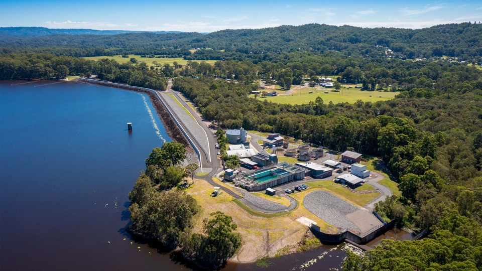 aerial view of a water treatment facility situated beside a large body of water, surrounded by dense green forested areas and rolling hills. The facility consists of several buildings, tanks, and infrastructure, positioned near a long, straight road running parallel to the water. The landscape includes a mix of forested and cleared areas with a few scattered buildings and open fields in the background.