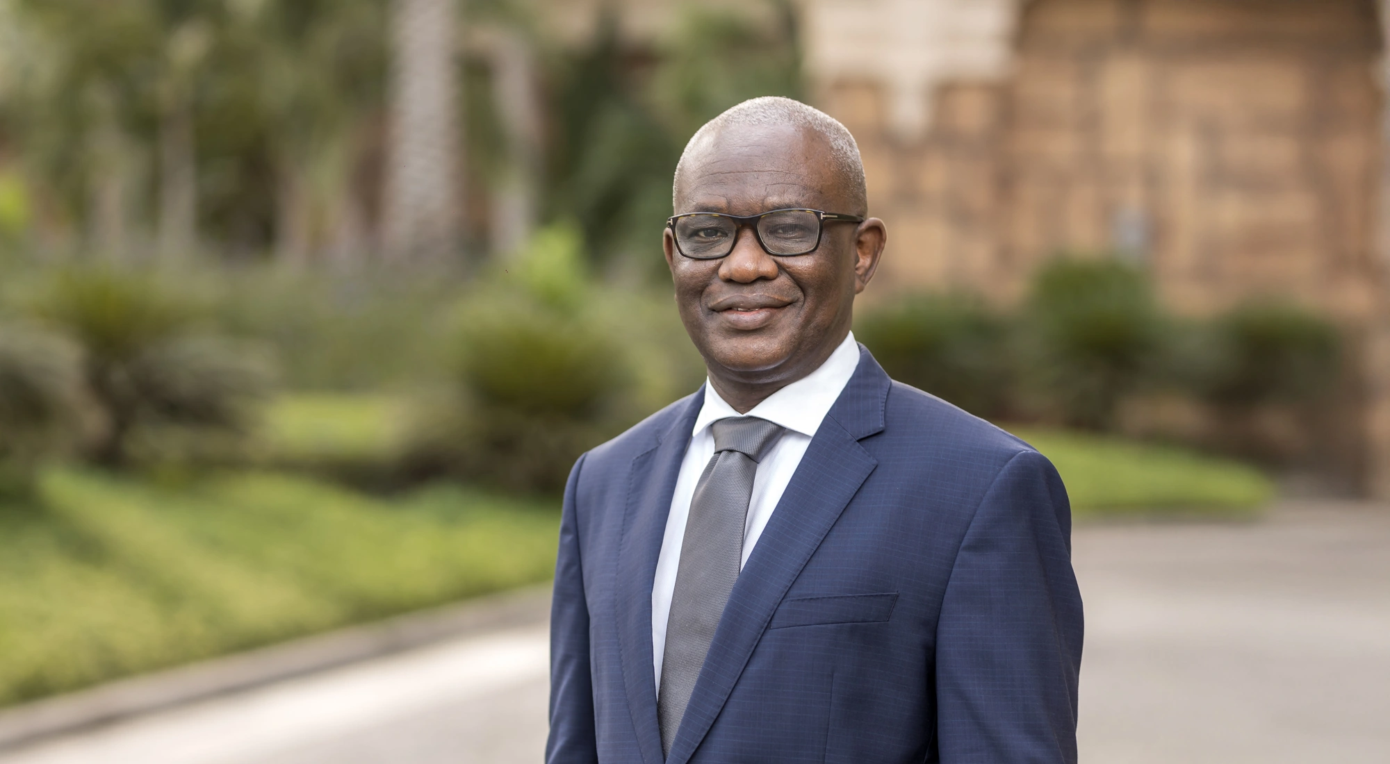 Portrait of Segun Faniran, Forensic Planning and Delay, Middle East, outside in front of grass and bushes with tall buildings behind.