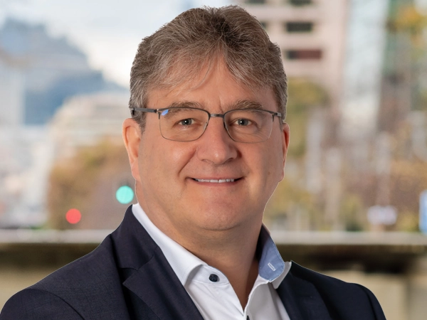 Portrait of Mathew Riley, Director, Aviation Lead, NAM, outside in front of a blurred brown background featuring a building and mix of natural and urban elements.