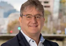 Portrait of Mathew Riley, Director, Aviation Lead, NAM, outside in front of a blurred brown background featuring a building and mix of natural and urban elements.