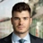 Portrait of Christian Weitz, Senior Project Manager, standing outside in front of buildings featuring an urban cityscape with a mix of architectural styles. The scene includes some windows and red features, adding a touch of color.