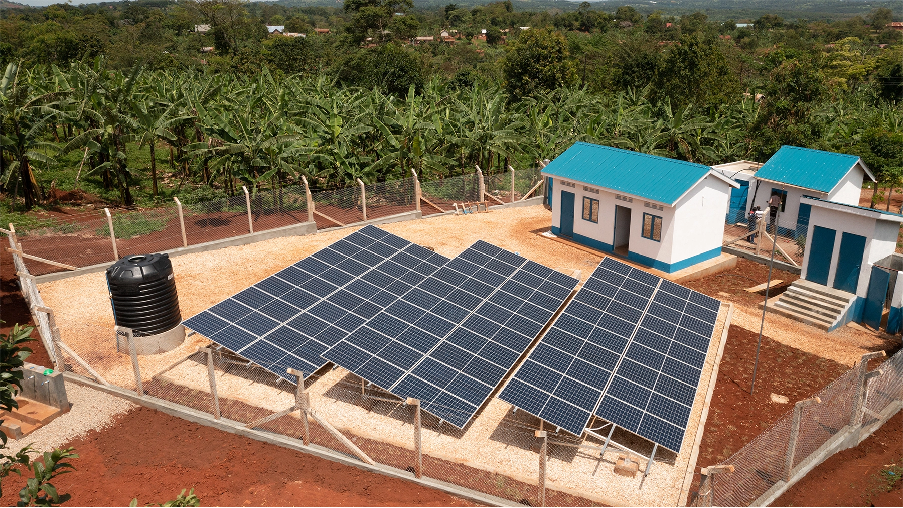 Three solar panels next to a few small houses with blue roofs, with a background of jungle foliage.