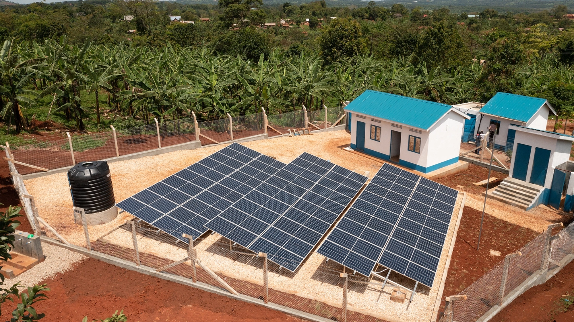 Three solar panels next to a few small houses with blue roofs, with a background of jungle foliage.